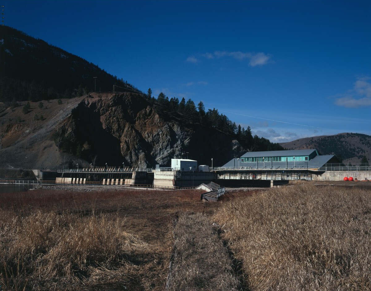 Milltown Dam's concrete spillway gates and powerhouse on the Clark Fork River in 2007, seen across dry grass below a rocky hillside